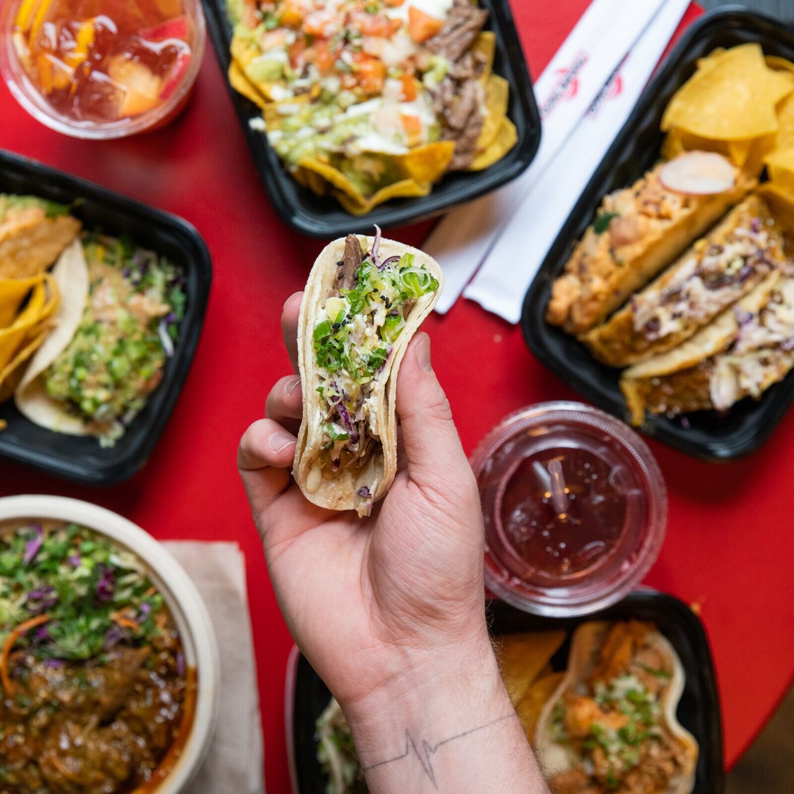 hand holding soft shell taco over red table with a spread of other takumi menu items 