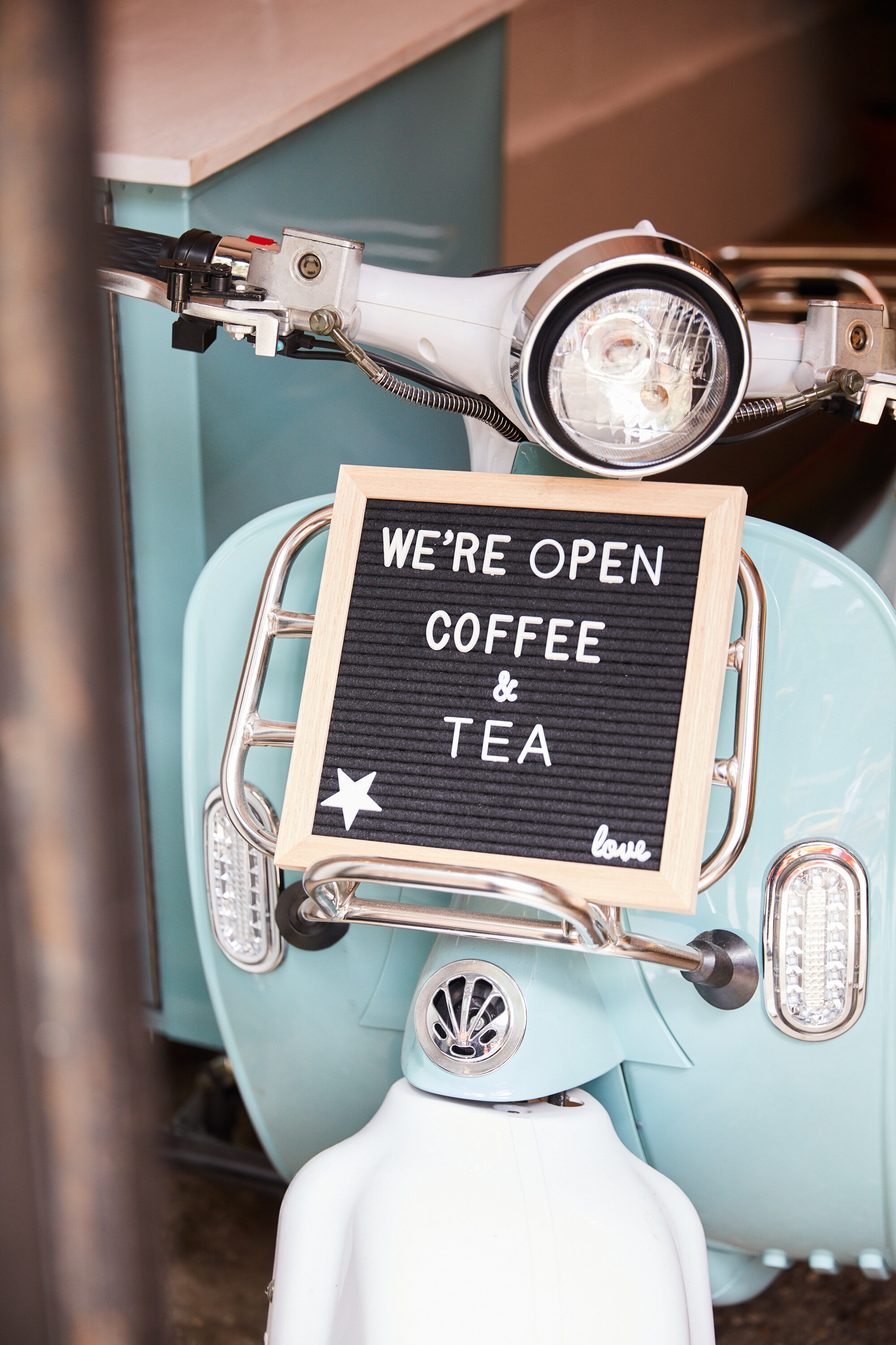  Blue Vespa with a letterboard on the from t that reads “we’re open. coffee and tea” 