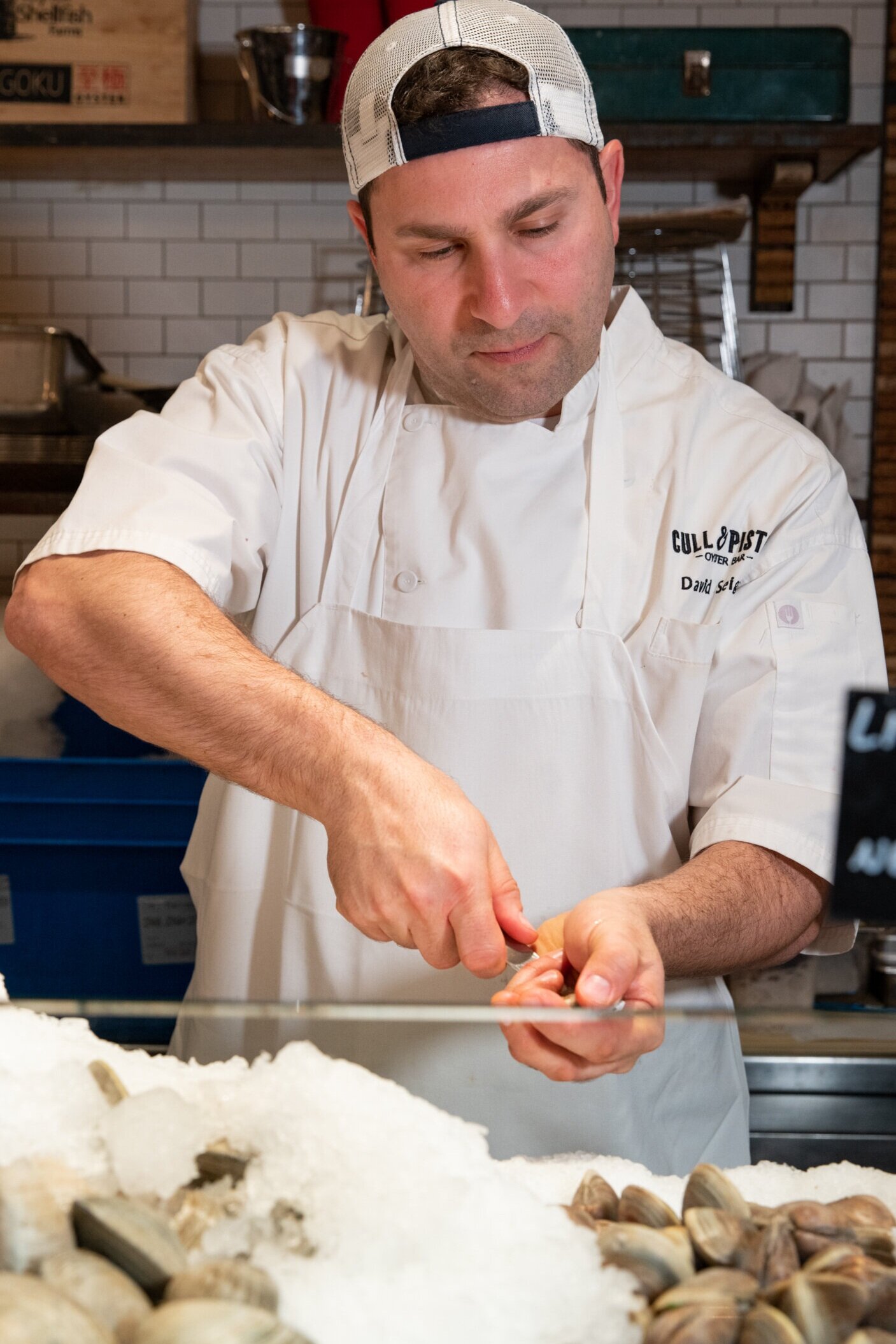 man shucking oyster wearing a hat and chefs coat reading cull and pistol