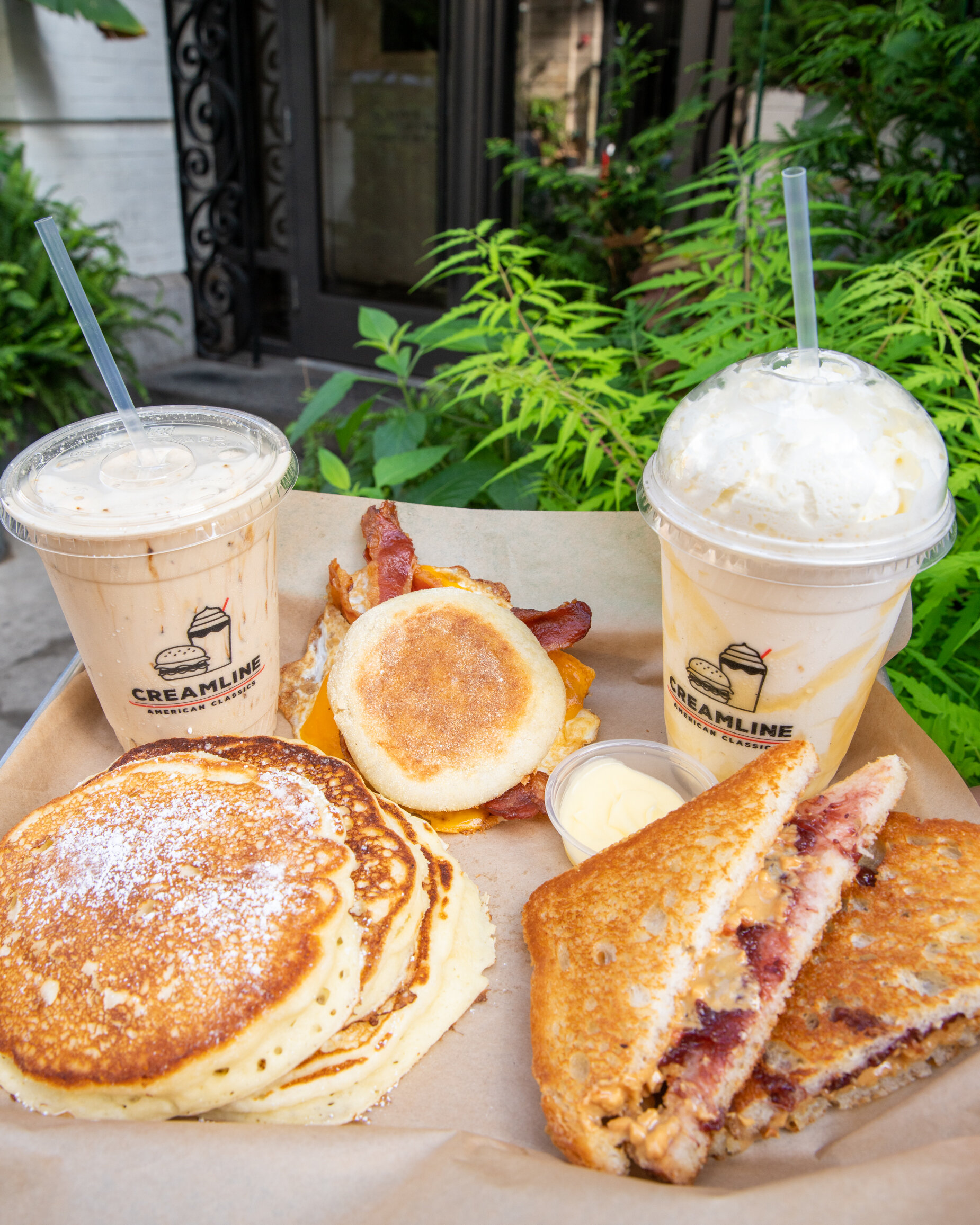 stack of pancakes, breakfast sandwich with bacon sticking out, peanut butter and jelly with two milkshakes 